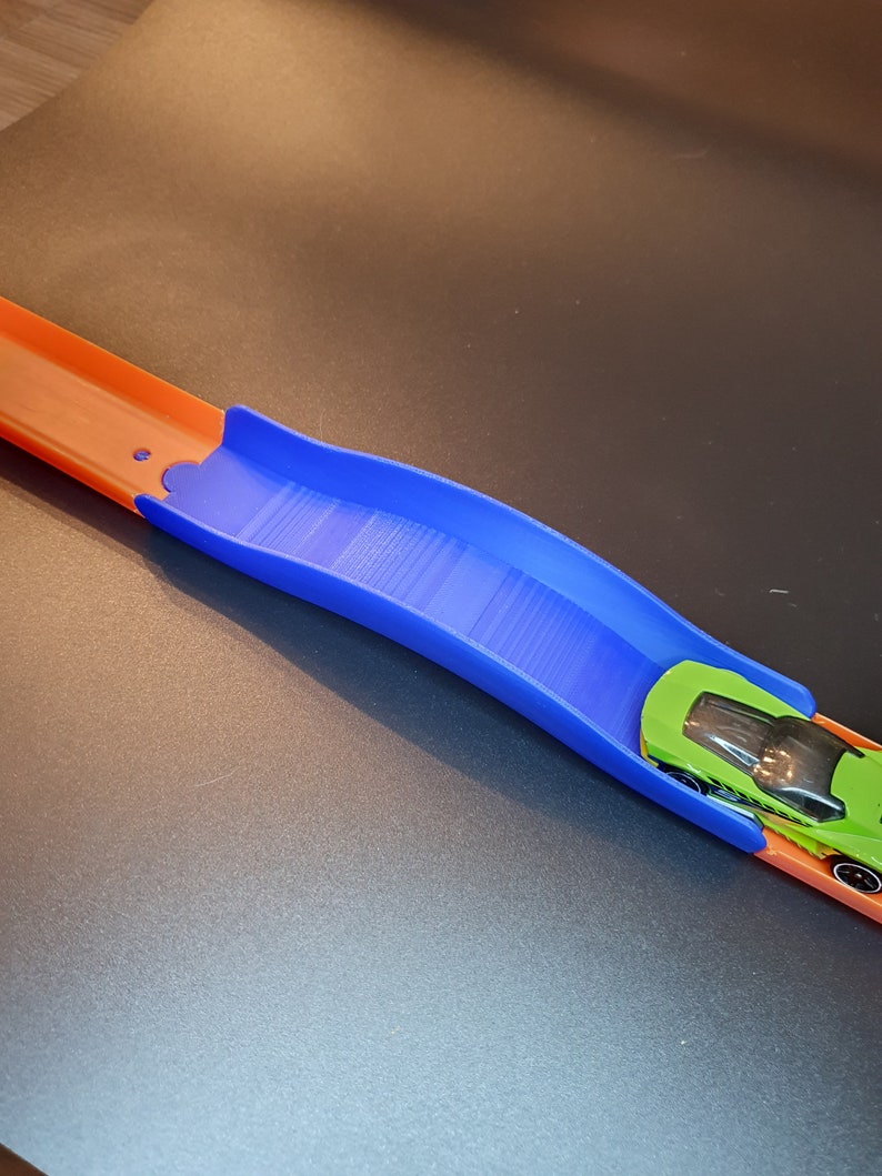 Op de afbeelding: Een blauw plastic baanstuk voor een speelgoedauto racebaan. Het baanstuk is gebogen en een groene speelgoedauto rijdt eroverheen.