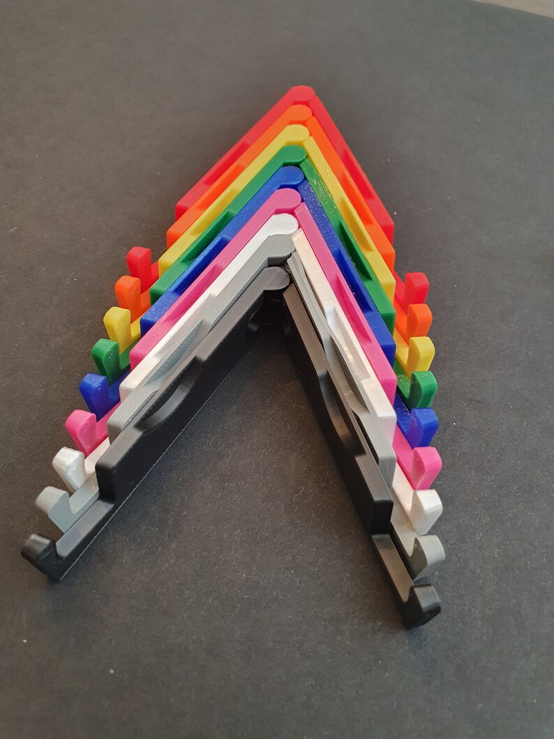 Puede incluir: Un conjunto de diez soportes para tel&eacute;fono de varios colores, incluyendo rojo, naranja, amarillo, verde, azul, morado, rosa, blanco, gris y negro. Los soportes est&aacute;n doblados en forma de V y apilados uno encima del otro.