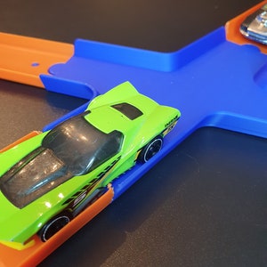May include: A green toy car with black wheels and yellow accents is on a blue and orange track. The car is on a blue section of the track and is about to go onto an orange section.