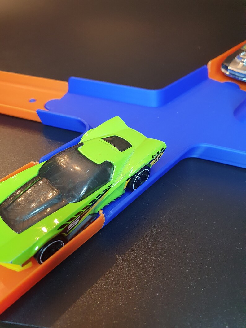 May include: A green toy car with black wheels and yellow accents is on a blue and orange track. The car is on a blue section of the track and is about to go onto an orange section.