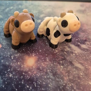 May include: Two small, 3D-printed cow figurines. One is brown with a white face, the other is white with black spots. Both have black hooves and horns. They are set against a galaxy-themed background.