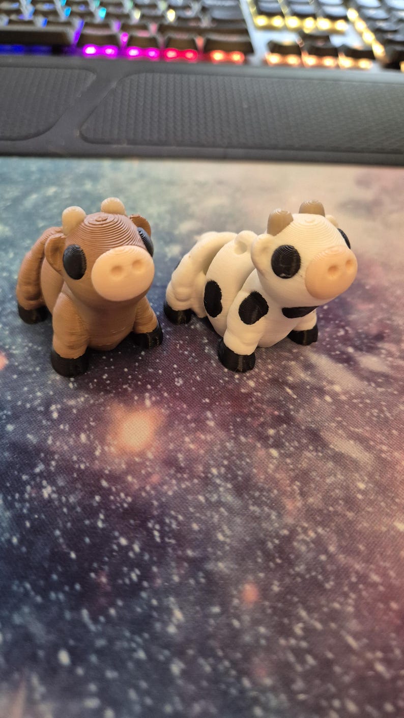 May include: Two small, 3D-printed cow figurines. One is brown with a white face, the other is white with black spots. Both have black hooves and horns. They are set against a galaxy-themed background.