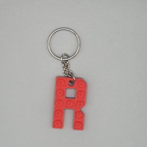 Peut inclure: Porte-cl&eacute;s en plastique rouge en forme de lettre R avec un anneau et une cha&icirc;ne en m&eacute;tal argent&eacute;. La lettre R est con&ccedil;ue pour ressembler &agrave; une brique Lego.