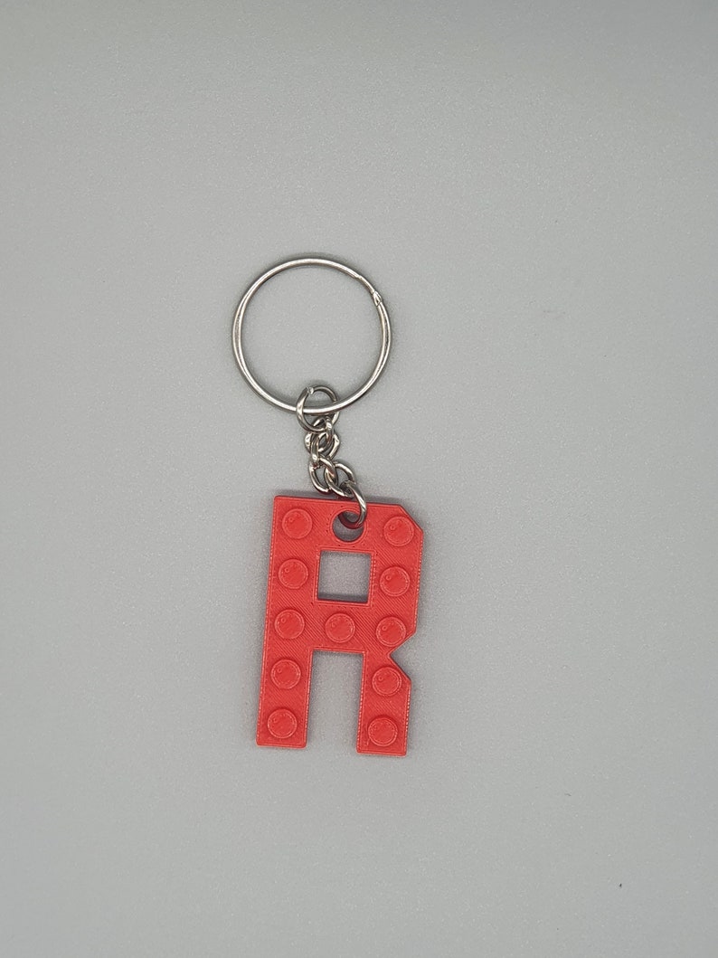 Peut inclure: Porte-cl&eacute;s en plastique rouge en forme de lettre R avec un anneau et une cha&icirc;ne en m&eacute;tal argent&eacute;. La lettre R est con&ccedil;ue pour ressembler &agrave; une brique Lego.