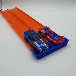May include: Two toy race cars, a blue and a red one, sit in a blue tray at the end of an orange race track. The blue car has the number 20 on it. The track is elevated and ready for racing.