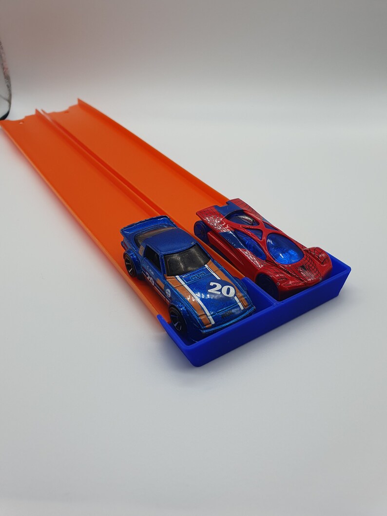 May include: Two toy race cars, a blue and a red one, sit in a blue tray at the end of an orange race track. The blue car has the number 20 on it. The track is elevated and ready for racing.