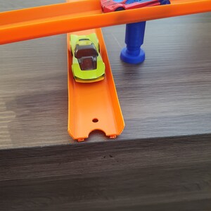Puede incluir: Dos coches de juguete en una pista de pl&aacute;stico naranja. La pista superior est&aacute; elevada y sostenida por un soporte de pl&aacute;stico azul. El coche de la pista superior es rojo con un parabrisas negro. El coche de la pista inferior es verde con un parabrisas negro.