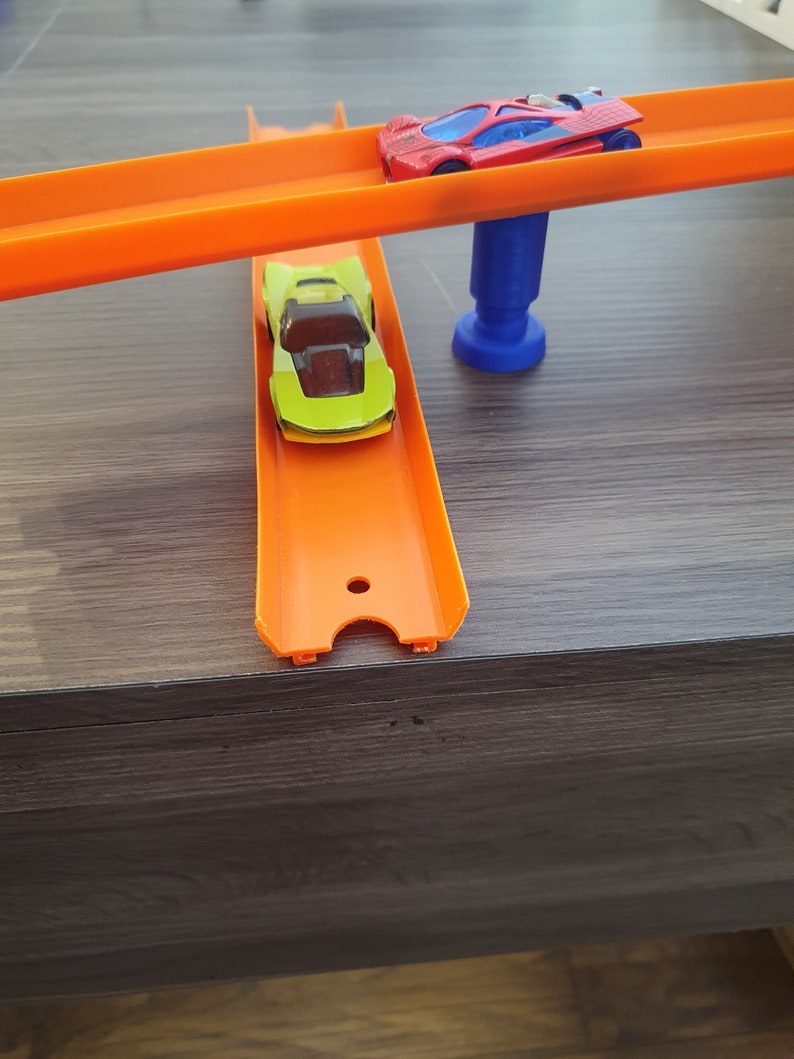 Puede incluir: Dos coches de juguete en una pista de pl&aacute;stico naranja. La pista superior est&aacute; elevada y sostenida por un soporte de pl&aacute;stico azul. El coche de la pista superior es rojo con un parabrisas negro. El coche de la pista inferior es verde con un parabrisas negro.