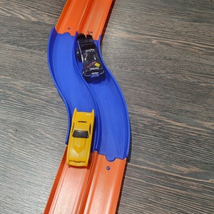 May include: Two toy cars race on a blue and orange track. The yellow car is in the lead, while the black and white police car is behind it.