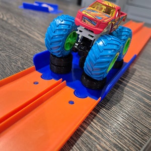 May include: A red monster truck toy with blue and green wheels is on an orange track. The truck is on a blue and orange ramp.