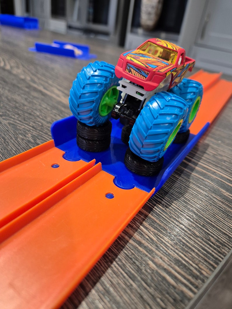 May include: A red monster truck toy with blue and green wheels is on an orange track. The truck is on a blue and orange ramp.