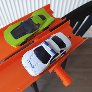 May include: Two toy cars on an orange track. The green car is in the background and the white police car is in the foreground. The police car has a blue light on top and the words "POLICE" printed on the hood.
