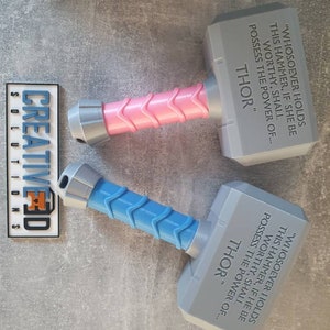May include: Two gray plastic toy hammers with blue and pink handles. The hammers have the inscription "Whosoever holds this hammer, if she be worthy, shall possess the power of... Thor" on them.