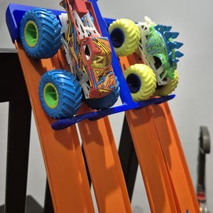 May include: Two toy monster trucks, one red and one green, are positioned on a Hot Wheels track. The red truck has a lightning bolt design and the green truck has a dinosaur design. The track is orange and blue and is attached to a blue plastic stand.