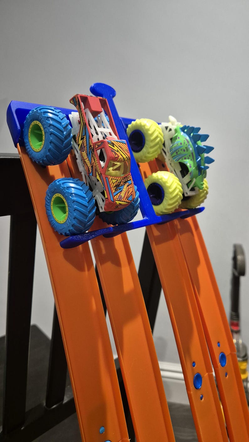 May include: Two toy monster trucks, one red and one green, are positioned on a Hot Wheels track. The red truck has a lightning bolt design and the green truck has a dinosaur design. The track is orange and blue and is attached to a blue plastic stand.