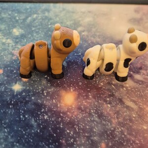 May include: Two articulated animal figures: a brown horse and a white cow with black spots. Both are 3D-printed and have segmented bodies, allowing for posing. They sit on a galaxy-themed surface, with a keyboard in the background.