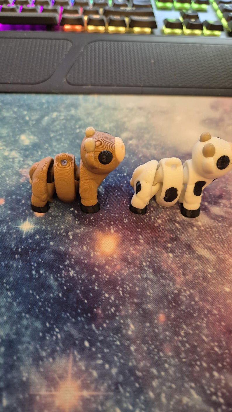 May include: Two articulated animal figures: a brown horse and a white cow with black spots. Both are 3D-printed and have segmented bodies, allowing for posing. They sit on a galaxy-themed surface, with a keyboard in the background.