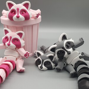 May include: Three 3D printed raccoon figurines. Two are pink and white, one is gray and white. One pink raccoon is in a pink trash can. The other two raccoons are lying down.