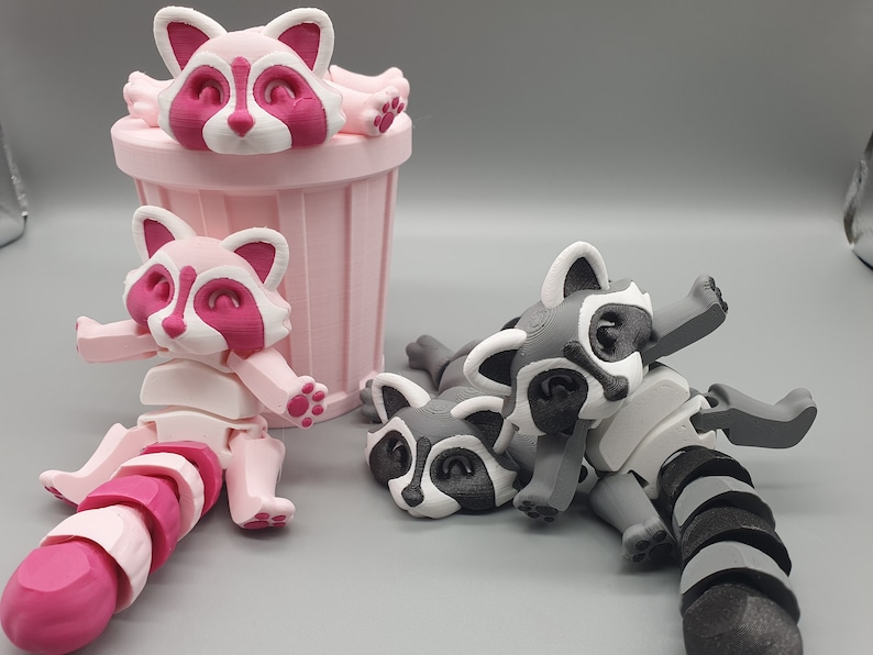 May include: Three 3D printed raccoon figurines. Two are pink and white, one is gray and white. One pink raccoon is in a pink trash can. The other two raccoons are lying down.