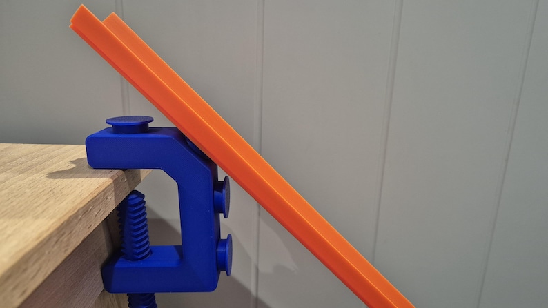 May include: A blue plastic clamp holding a bright orange track piece. The clamp is attached to a wooden surface.