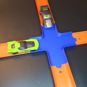 May include: A green and black toy car on an orange track with a blue connector piece. A black toy car is on the orange track above the connector piece.