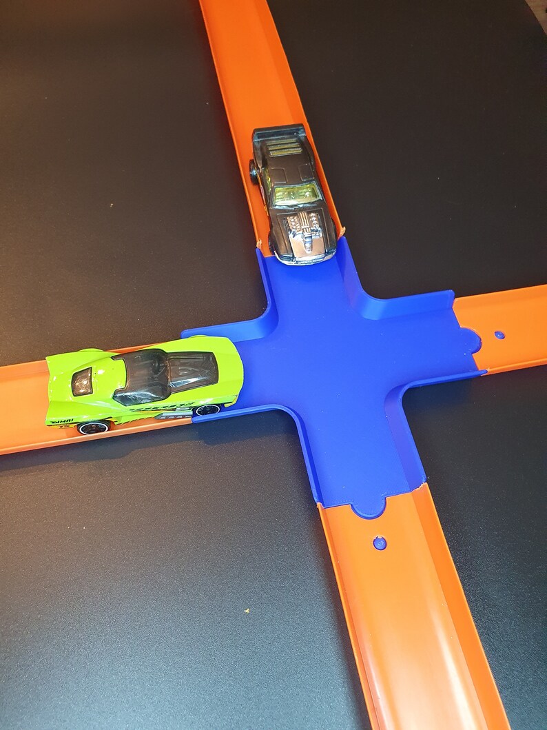 May include: A green and black toy car on an orange track with a blue connector piece. A black toy car is on the orange track above the connector piece.