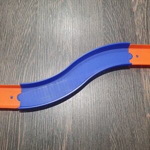 May include: A blue and orange plastic track piece for a toy car racing set. The track is curved and has two sections that connect together.