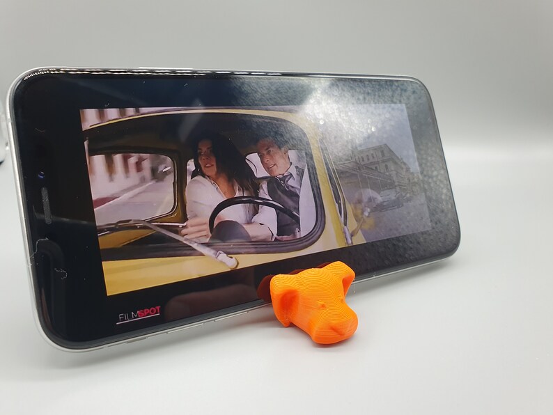 May include: A 3D printed orange dog head phone stand with a smartphone displaying a scene from a movie. The phone screen shows a couple driving in a yellow car. The text "FILMSPOT" is visible on the phone screen.