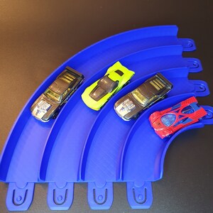 May include: Four toy cars on a blue plastic track. The cars are black, green, red, and black. The track is curved and has four sections.