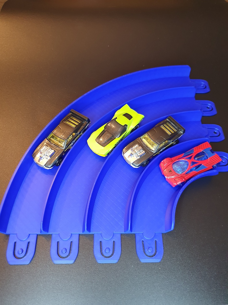 May include: Four toy cars on a blue plastic track. The cars are black, green, red, and black. The track is curved and has four sections.