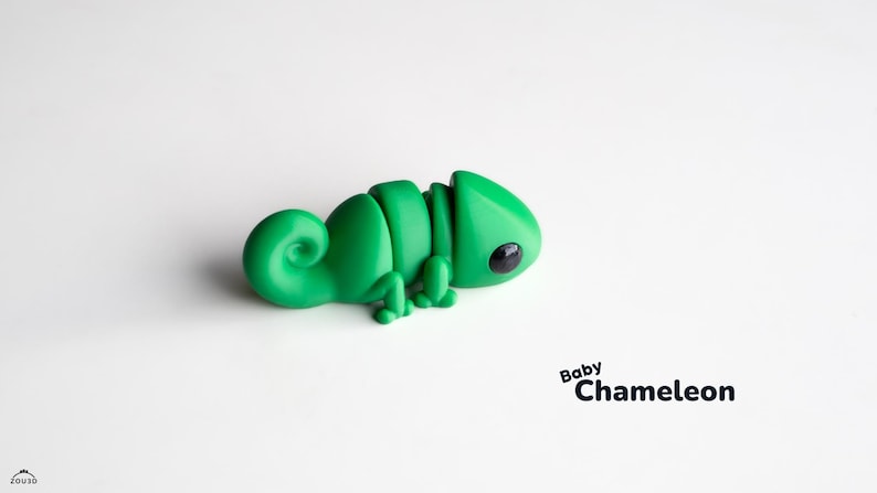 Op de afbeelding: Een groen, gesegmenteerd, 3D-geprint chameleonsculptuur met zwarte ogen. Het beeldje is geposeerd met zijn staart gekruld en zijn hoofd lichtjes gekanteld. De tekst "Baby Chameleon" is onder het beeldje gedrukt.