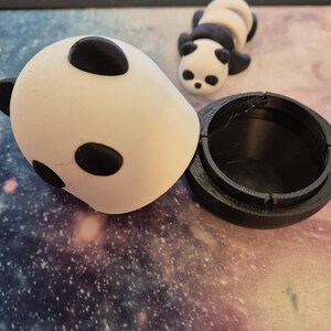 May include: A white and black panda-shaped container with a matching panda figurine. The container is open, revealing a black interior. The panda has black eyes and ears. The items are on a galaxy-themed surface.