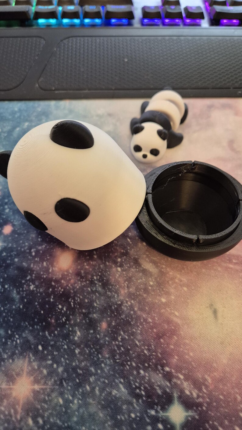 May include: A white and black panda-shaped container with a matching panda figurine. The container is open, revealing a black interior. The panda has black eyes and ears. The items are on a galaxy-themed surface.