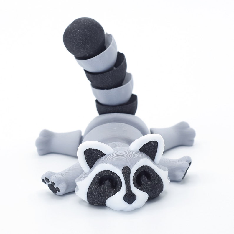 May include: A gray and black raccoon figurine with a segmented tail. The raccoon is lying on its belly with its head turned to the side and its eyes closed. The figurine is made of a 3D printed material.