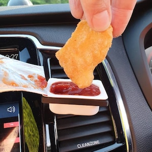 Pu&ograve; includere: Un porta condimento in plastica bianca con salsa rossa al suo interno. Una mano sta immergendo un bocconcino di pollo croccante e dorato nella salsa. Il porta condimento &egrave; attaccato alla bocchetta di aerazione di un'auto.