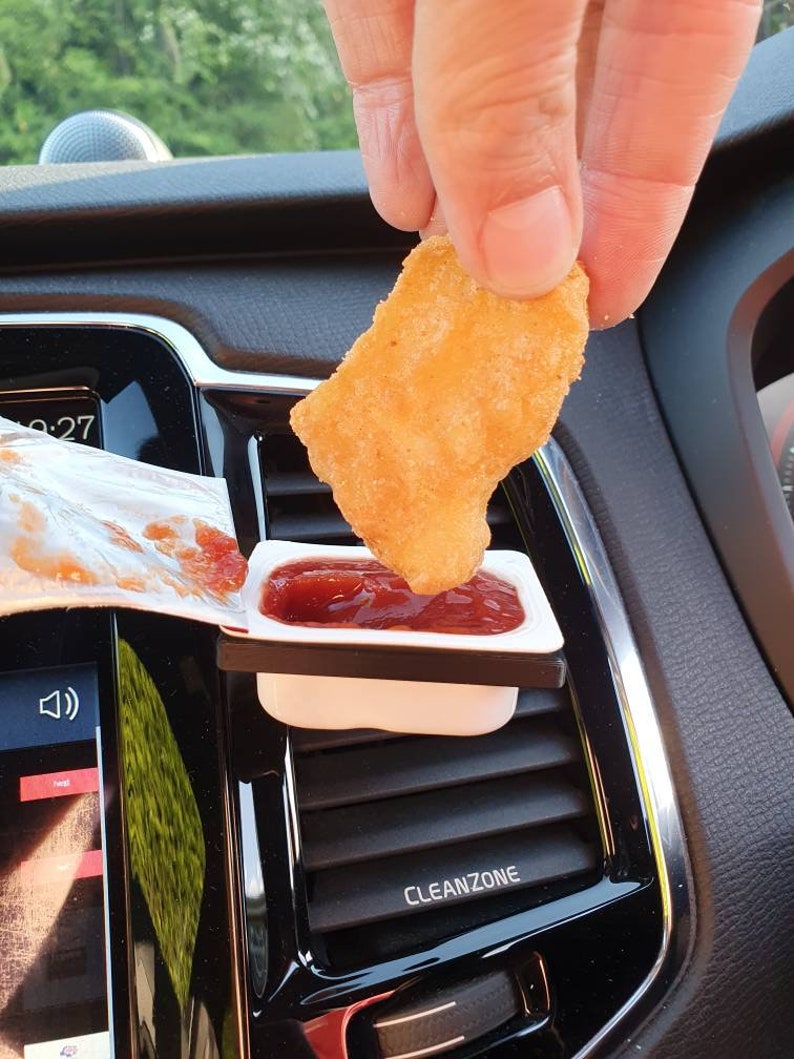 Pu&ograve; includere: Un porta condimento in plastica bianca con salsa rossa al suo interno. Una mano sta immergendo un bocconcino di pollo croccante e dorato nella salsa. Il porta condimento &egrave; attaccato alla bocchetta di aerazione di un'auto.