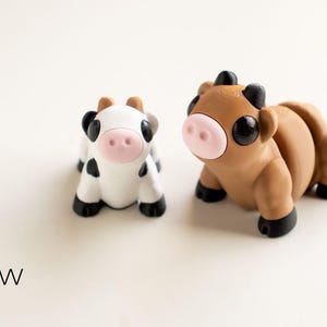 May include: Two miniature cow figurines. One is white with black spots, the other is brown. Both have pink noses, black hooves, and large black eyes. The text "Tiny Cow" is at the bottom.