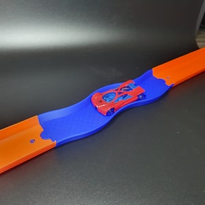 May include: A red toy car on a blue and orange plastic track. The track is curved and the car is in the middle of the curve.