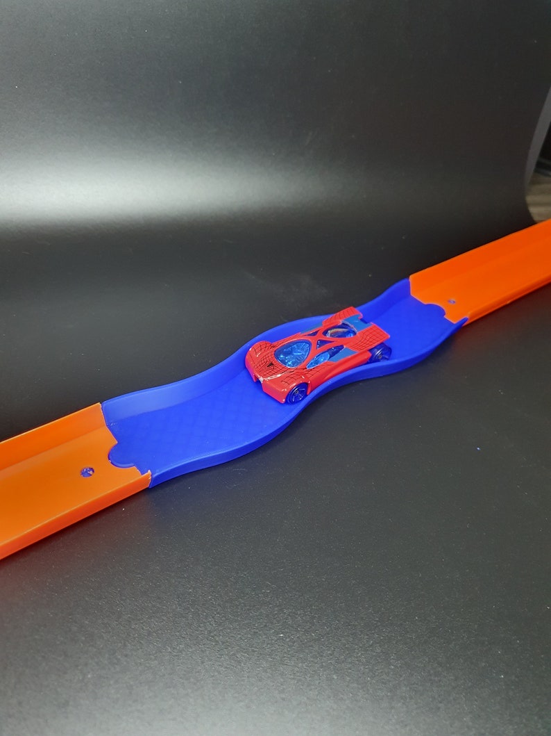 May include: A red toy car on a blue and orange plastic track. The track is curved and the car is in the middle of the curve.