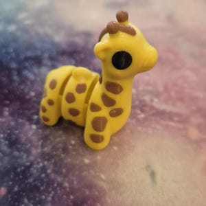 May include: A small yellow giraffe figurine with brown spots. The giraffe has a long neck and a small head with large black eyes.