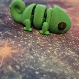 Op de afbeelding: Een groen 3D-geprint chameleonsculptuur met een zwart oog. Het beeldje staat op een kleurrijke stoffen achtergrond.