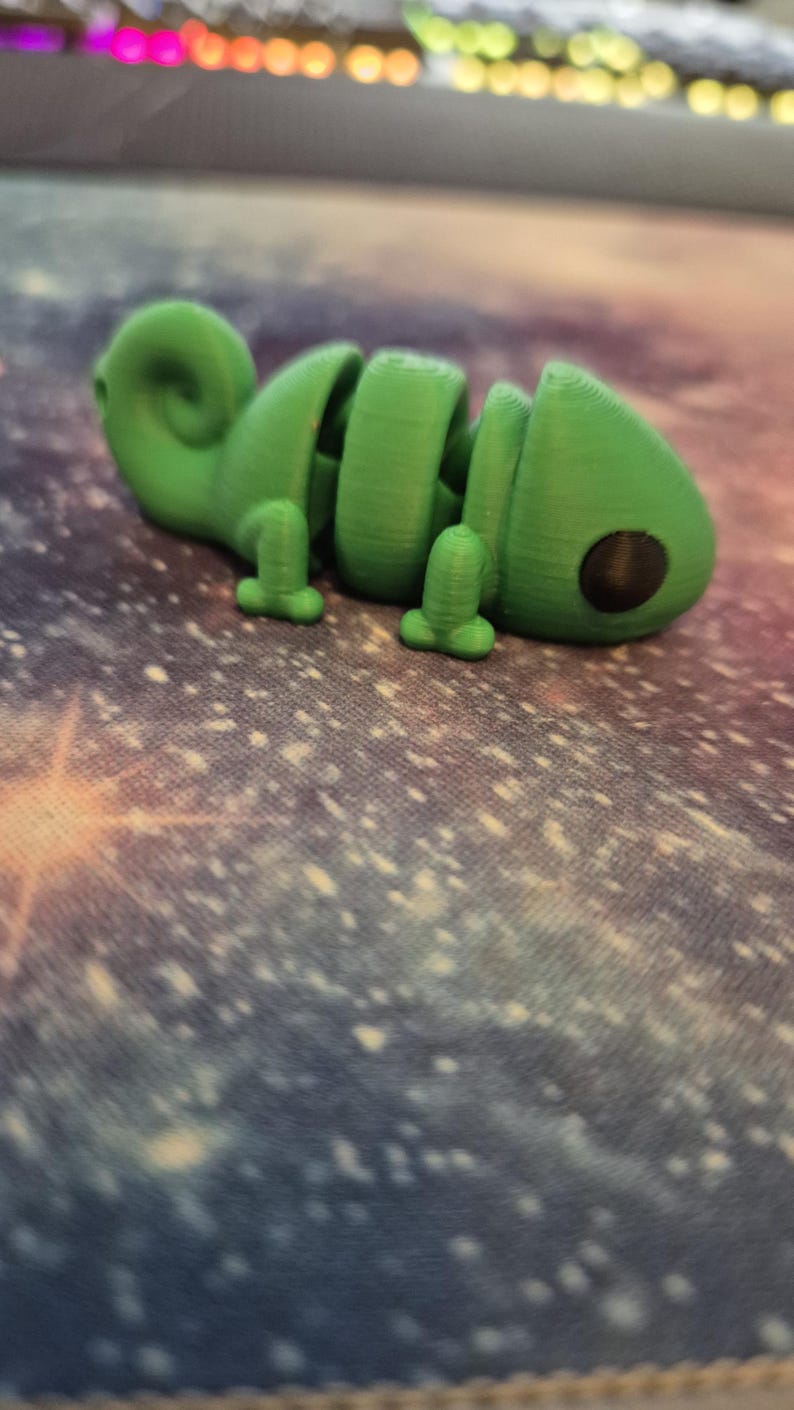 Op de afbeelding: Een groen 3D-geprint chameleonsculptuur met een zwart oog. Het beeldje staat op een kleurrijke stoffen achtergrond.