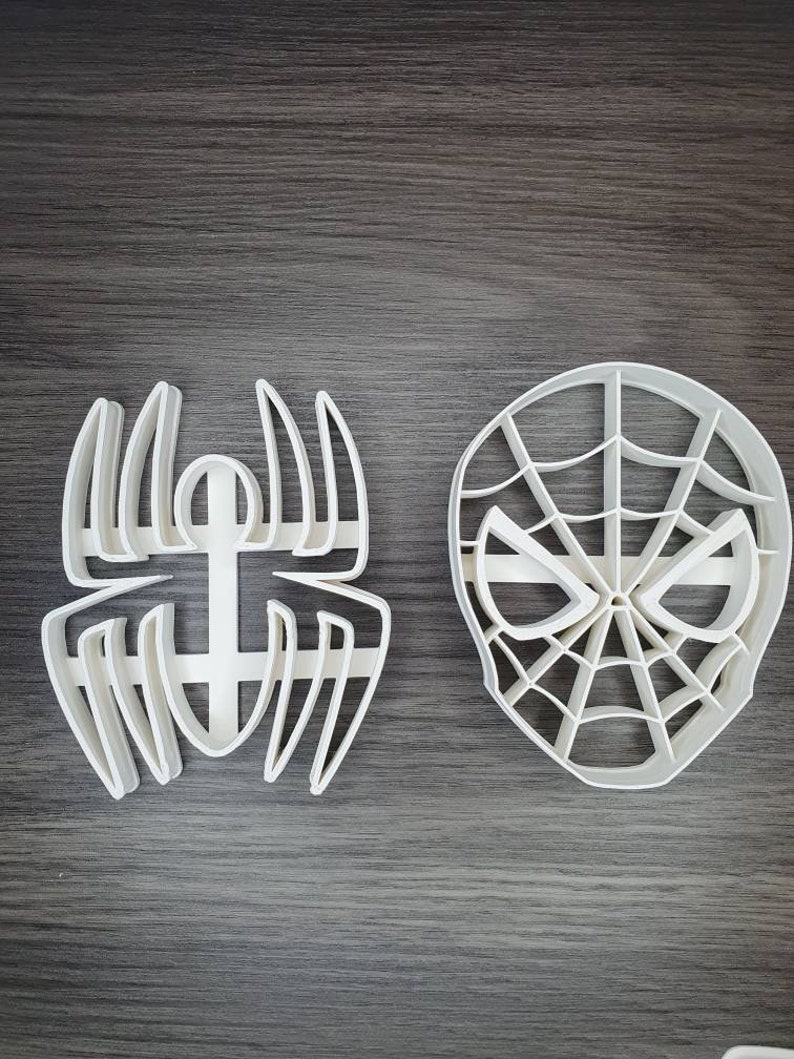May include: Two white plastic cookie cutters, one shaped like a spider and the other shaped like a Spiderman mask.