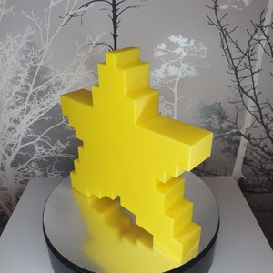 Puede incluir: Una escultura amarilla en forma de estrella pixelada sobre una base giratoria negra y plateada.