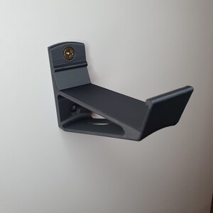 May include: A black plastic wall mount for a skateboard. The mount is attached to a white wall with a screw.