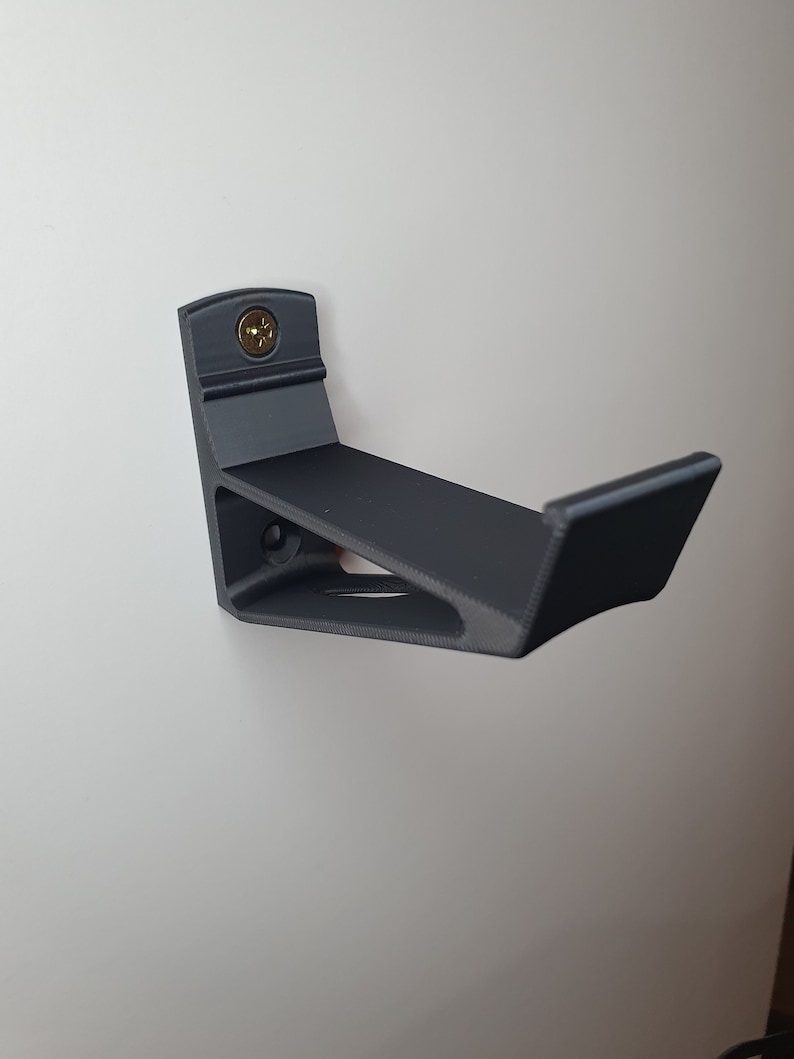 May include: A black plastic wall mount for a skateboard. The mount is attached to a white wall with a screw.