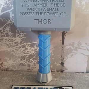 May include: A gray 3D printed replica of Thor's hammer, Mjolnir, with the inscription "WHOSOEVER HOLDS THIS HAMMER, IF HE BE WORTHY, SHALL POSSESS THE POWER OF... THOR" on the top. The hammer has a blue handle.