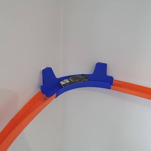 May include: A blue and orange plastic toy race track set up in a corner of a room. The track is curved and has a small toy car on it.