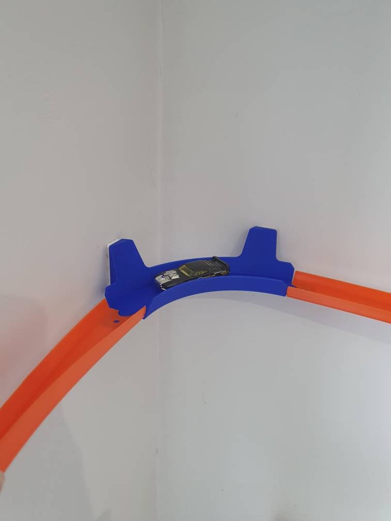 May include: A blue and orange plastic toy race track set up in a corner of a room. The track is curved and has a small toy car on it.