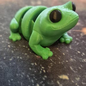 May include: A 3D printed green frog figurine with black eyes. The frog is sitting on a dark fabric surface with a speckled pattern.